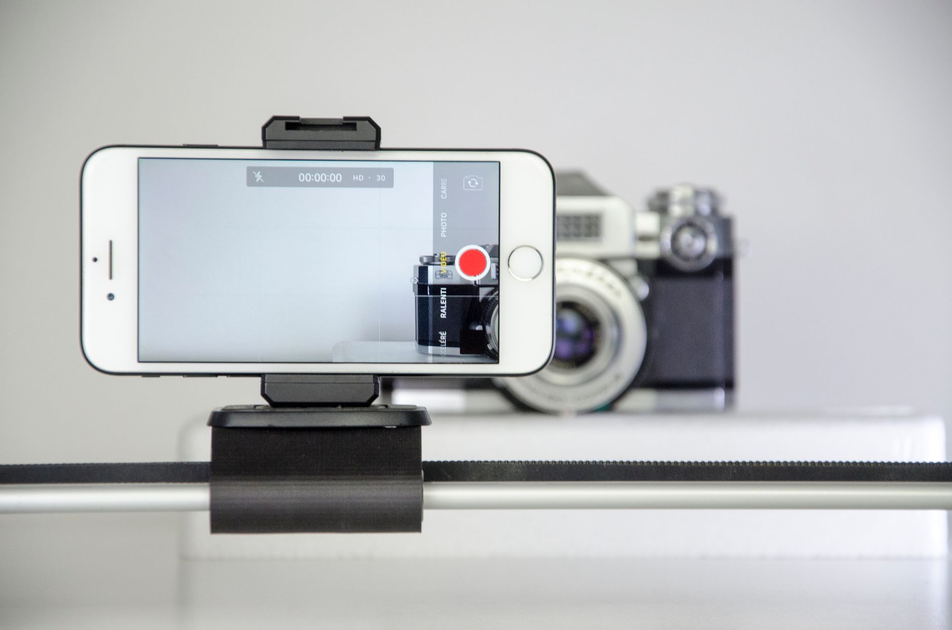 Gros plan sur un smartphone en mode vidéo, fixé sur un support, devant un appareil photo argentique flou.