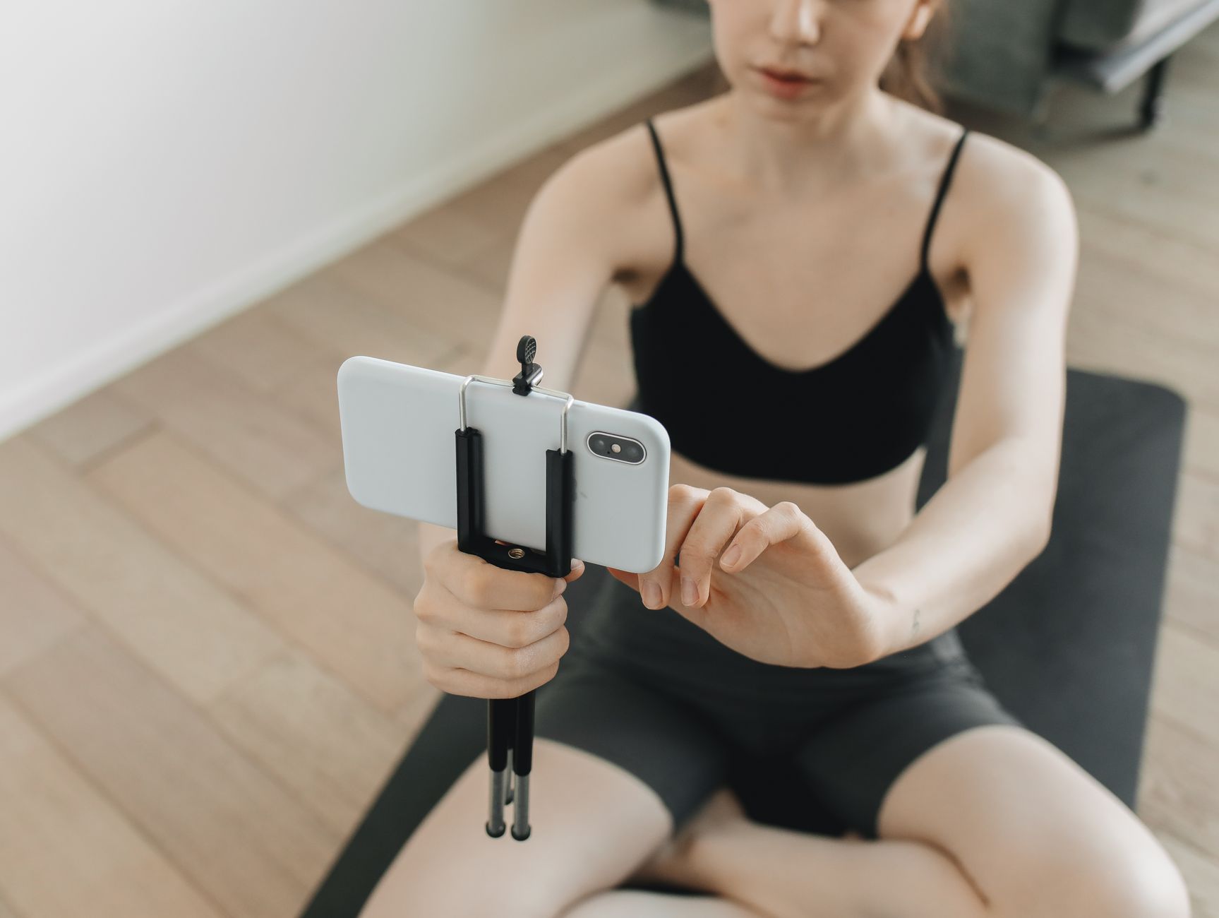 Une femme assise sur un tapis de sport dans son salon règle un smartphone fixé sur un petit trépied pour se filmer.